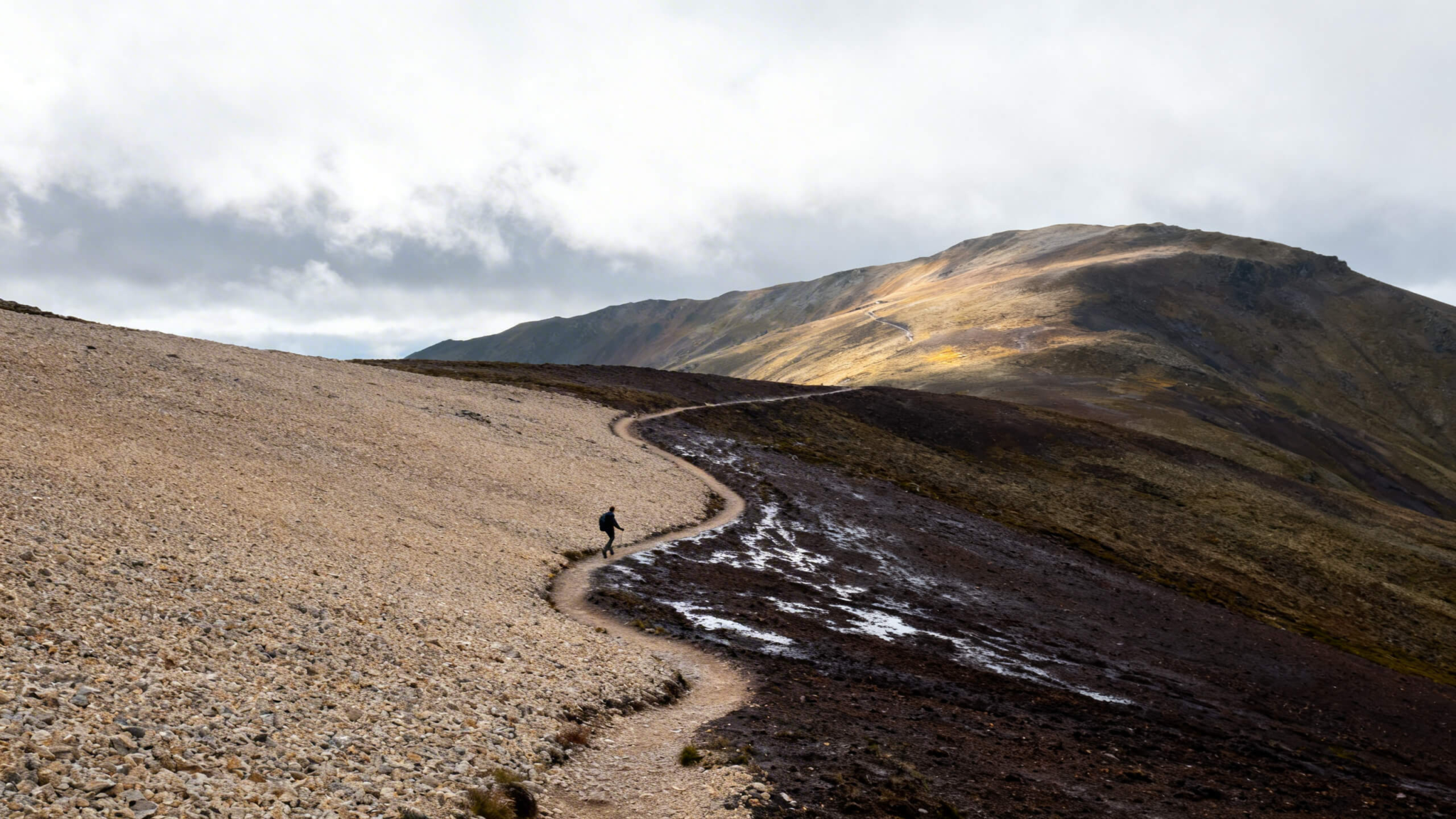 Mountain trail transitioning from dry to damp terrain under moving clouds with a lone hiker adapting stride.