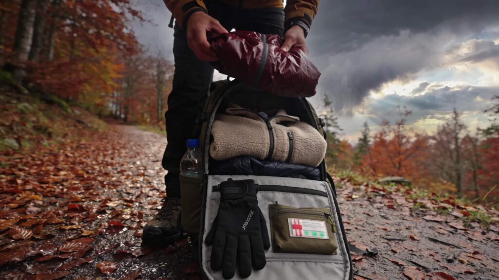 Hiker pulling a rain shell from a daypack on a leaf-covered trail, showing why changing weather requires a different backpack setup.