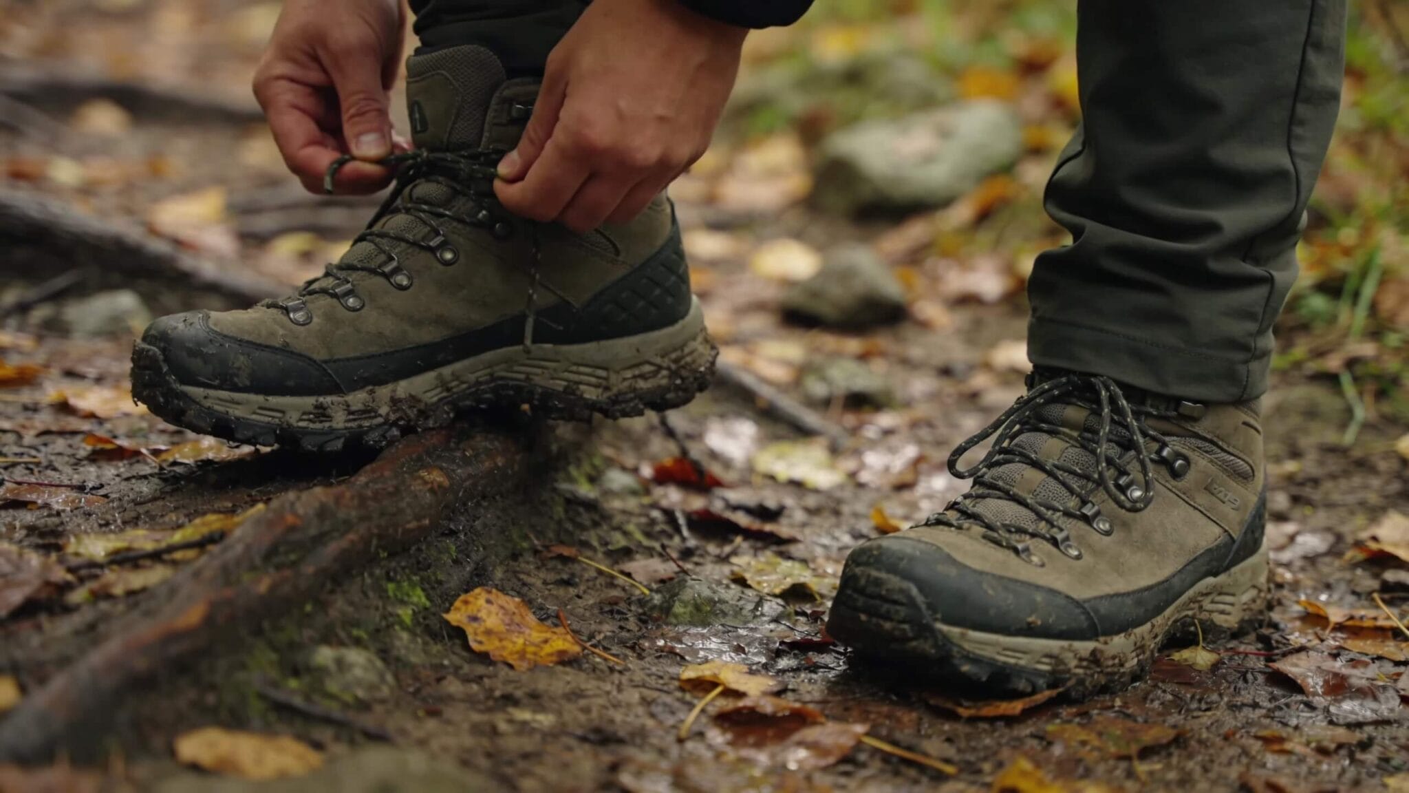 Hiker tying fall hiking boots on a wet leafy trail with mud, roots, and rocks, showing traction, support, and waterproof footwear for beginner hikers